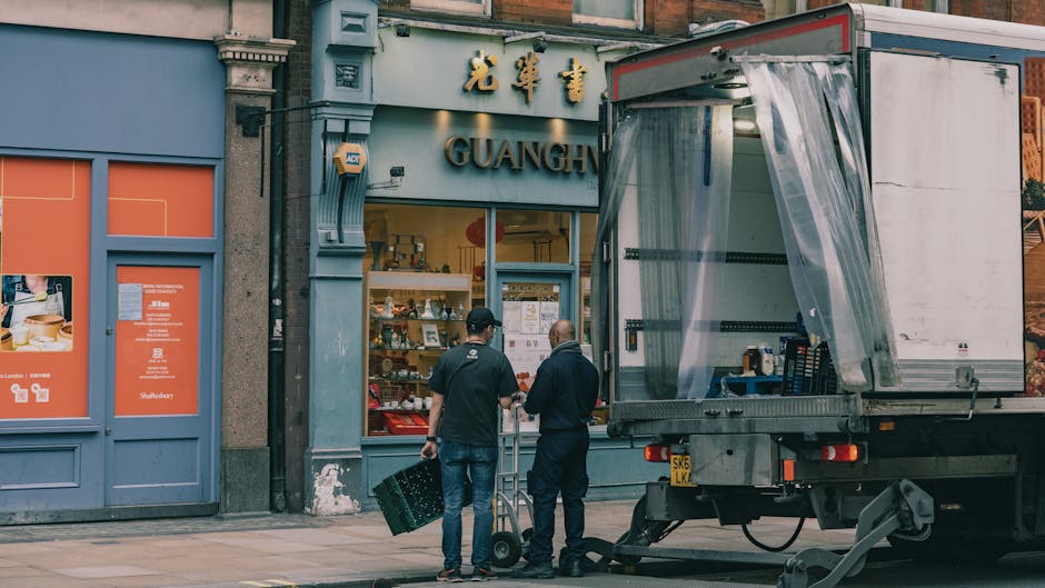A busy street scene in Soho, London, featuring a narrow pedestrianised street with multi-storey buildings on either side, some with decorative facades and large windows. People are walking along the s